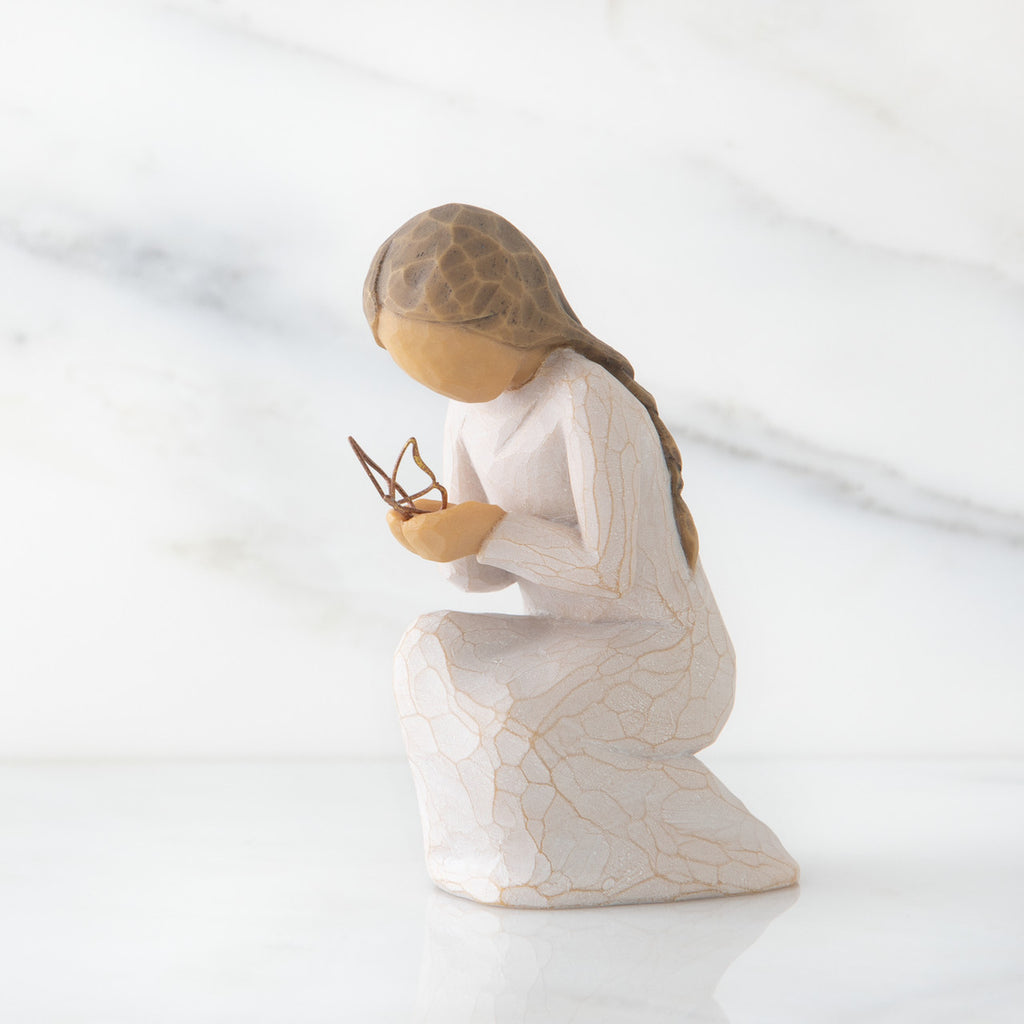 Side view of kneeling female figure in cream dress, with wire butterfly on extended hands, on marble background