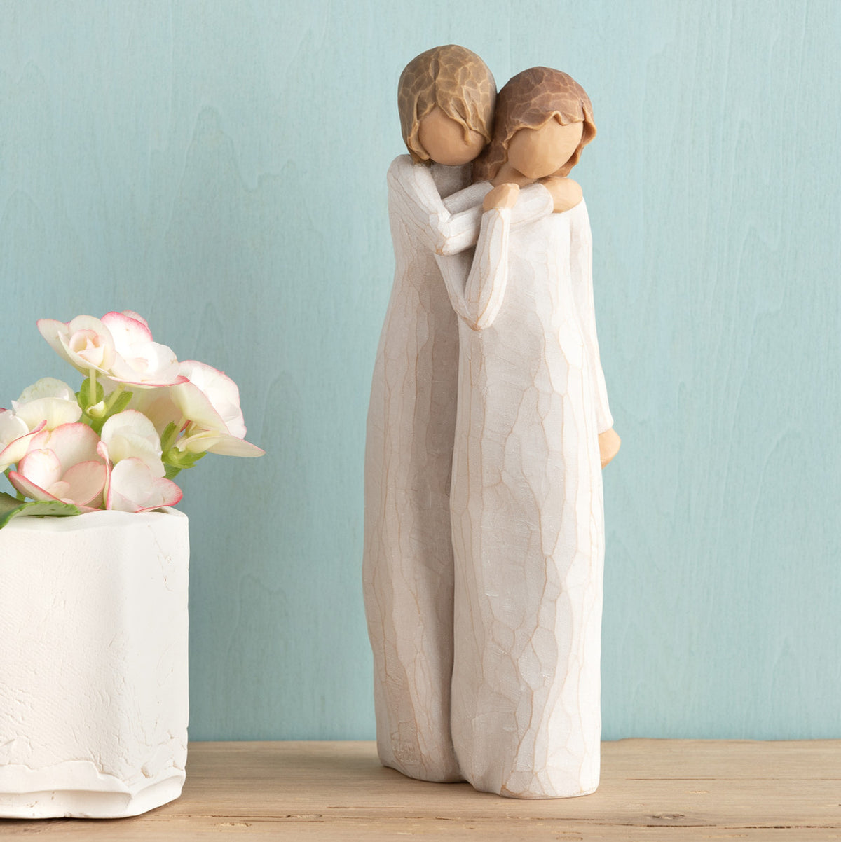Willow Tree Chrysalis, 9”h hand-painted standing resin figure of two women in cream dresses; one standing behind and embracing the other, on wooden shelf with flowers