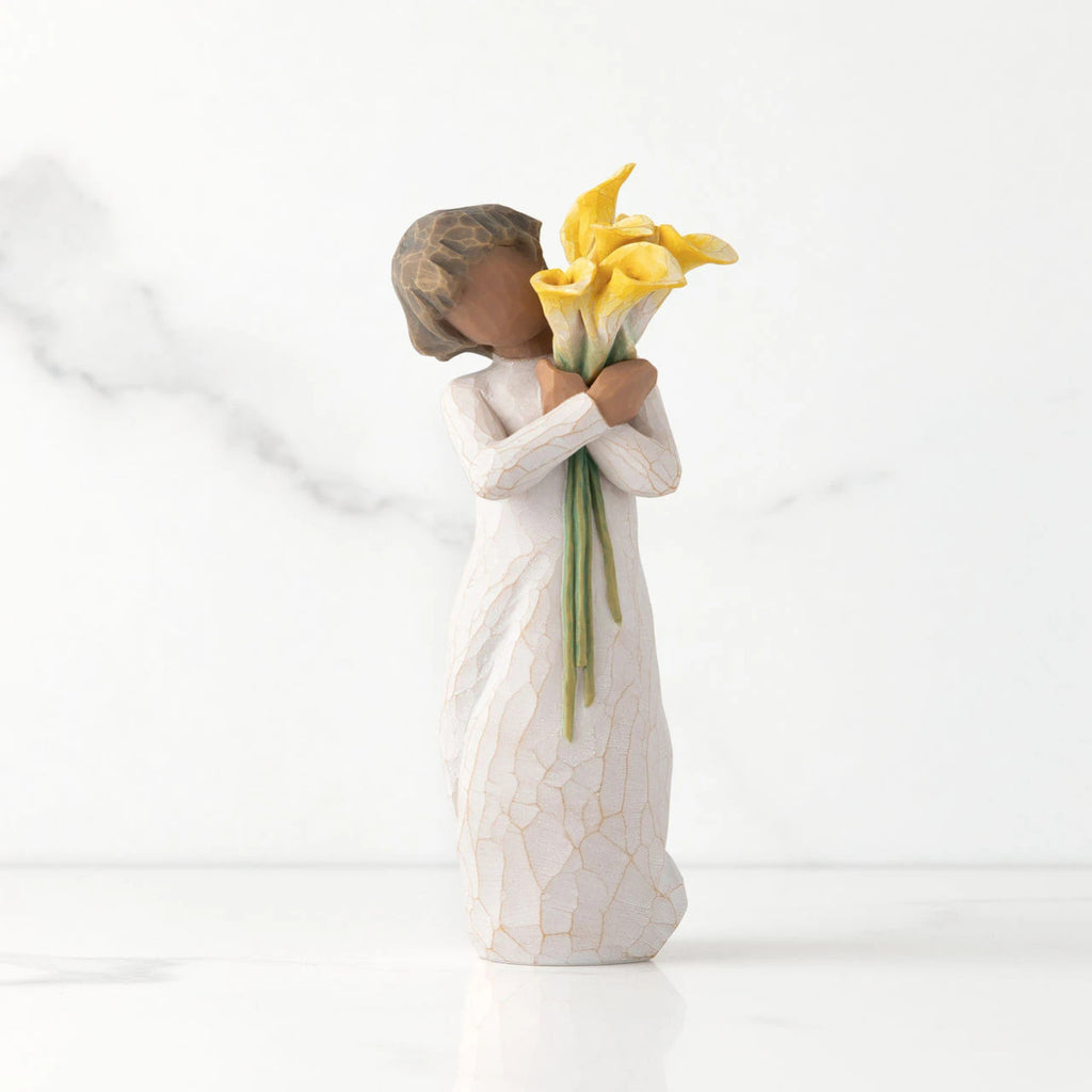 Front view of standing figure in cream dress with darker skin holding yellow calla lilies in her arms