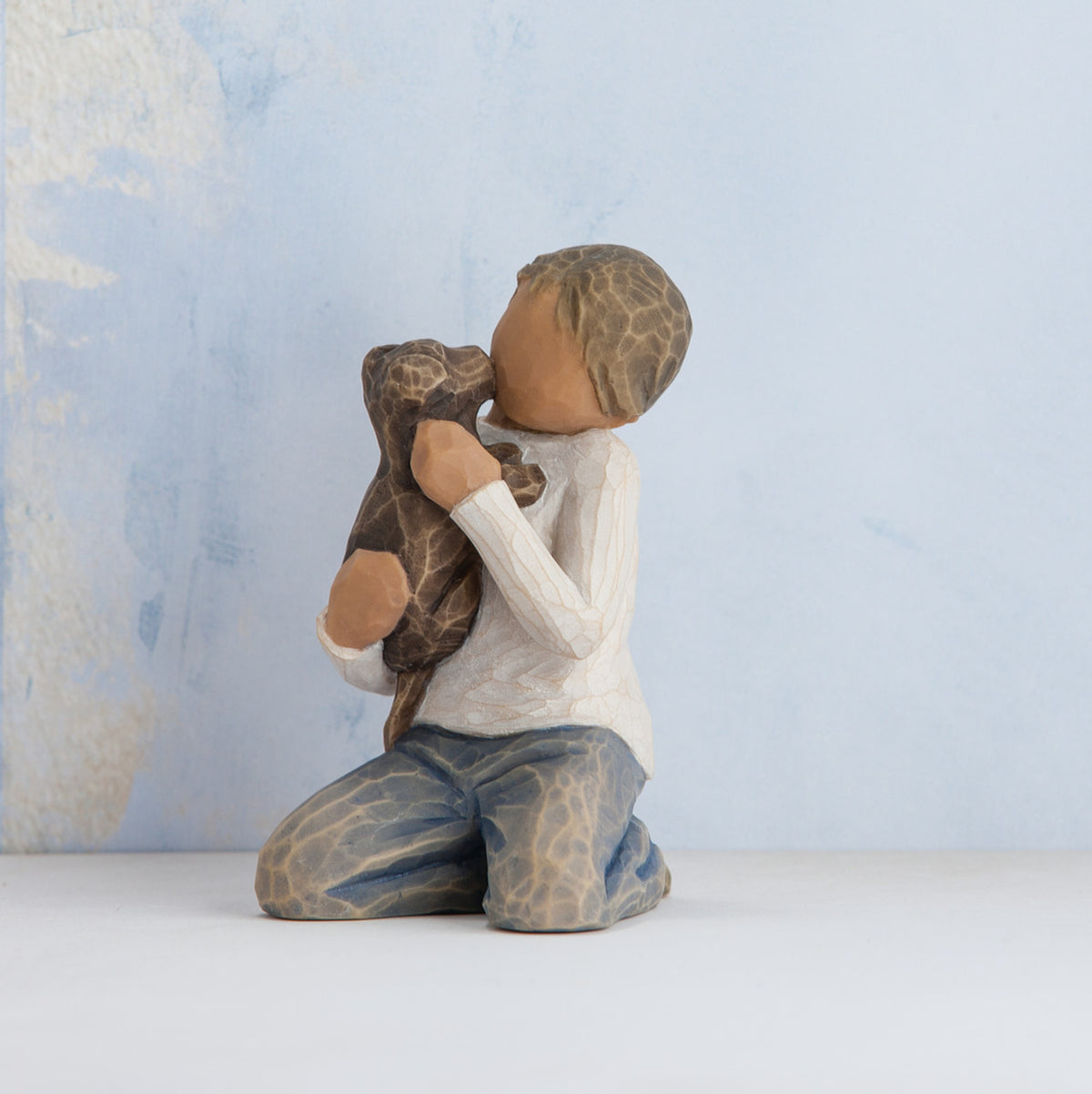 Willow Tree Kindness (boy), 3" hand-painted resin figure in neutral clothes, darker skin and hair, kneeling and holding black dog, in front of pale blue painting