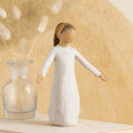Willow Tree Blessings, 5.5”h hand-painted resin figure in cream dress with darker skin and hair, wearing glitter headband, arms stretched out to sides, on table with pussywillows