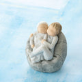 Willow Tree My new baby (sky), 2.5”h hand-painted resin figure of toddler in blue onesie holding infant in cream, cuddled together in a beanbag, in front of blue painting