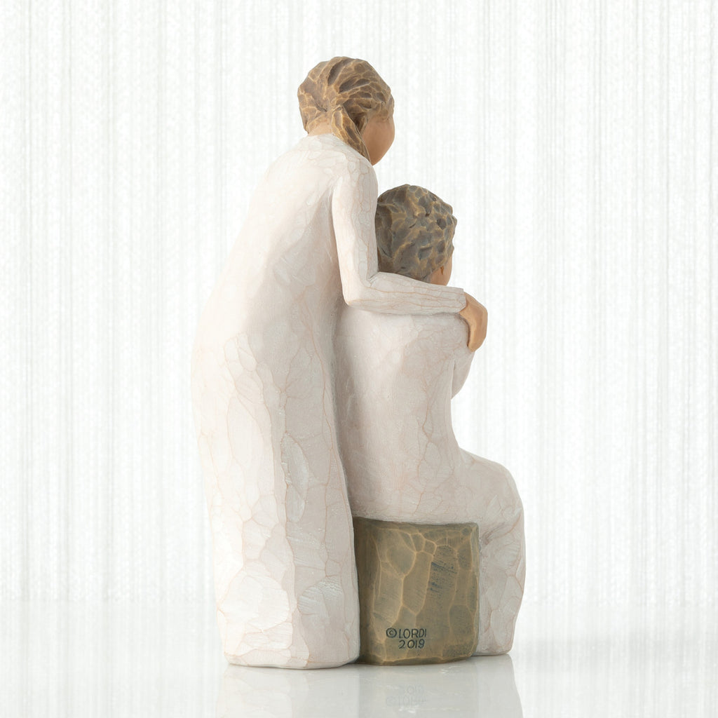 Back view, seated older female in cream dress, with younger female in cream dress, standing at her shoulder, hands embracing
