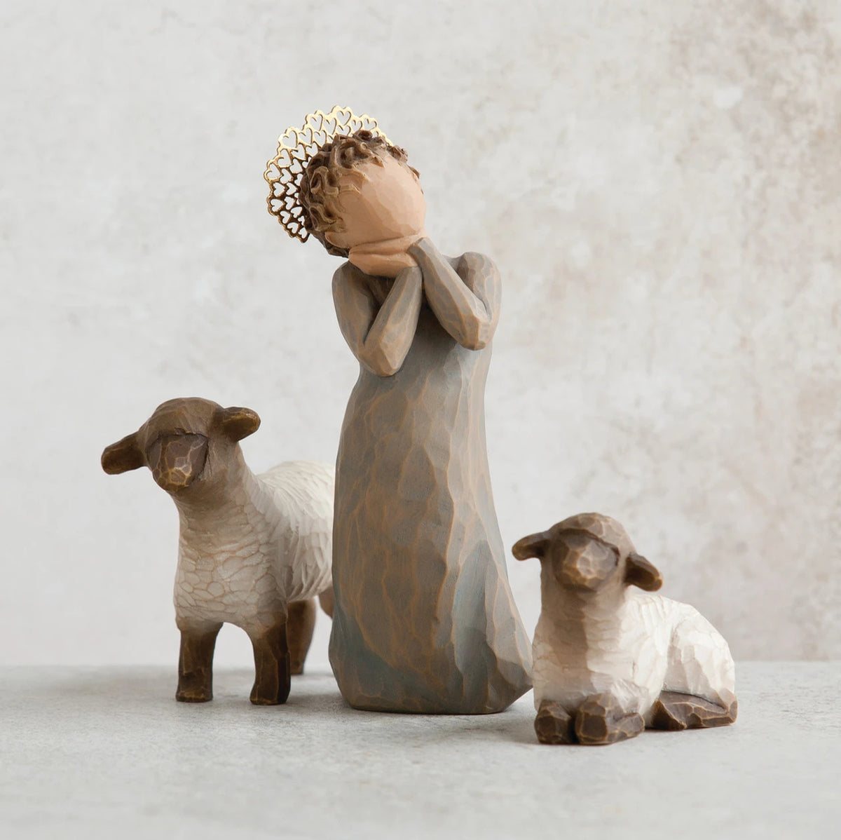 Close view of small angel figurine in dark tan dress holding hands up to face, in between to white/brown lamb figurines