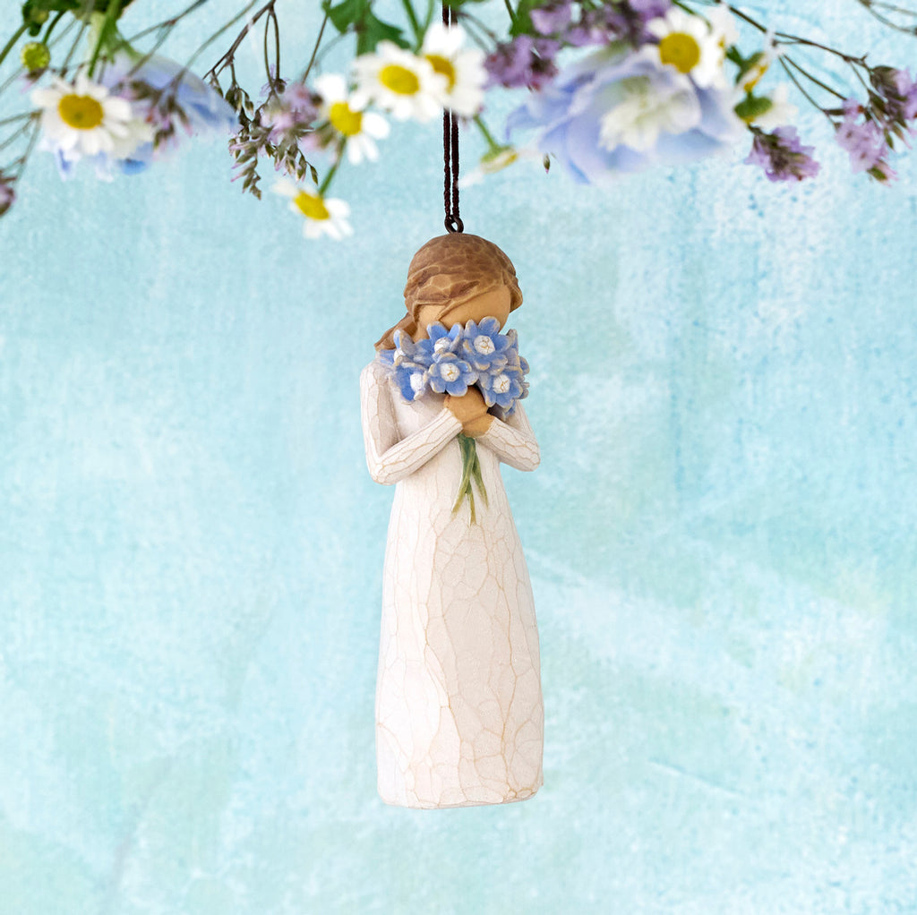 Willow Tree Forget-me-not Ornament, 4.25”h hand-painted resin figure in cream dress, holding bouquet blue flowers up to face, brown elastic cord, hanging in front of blue painting