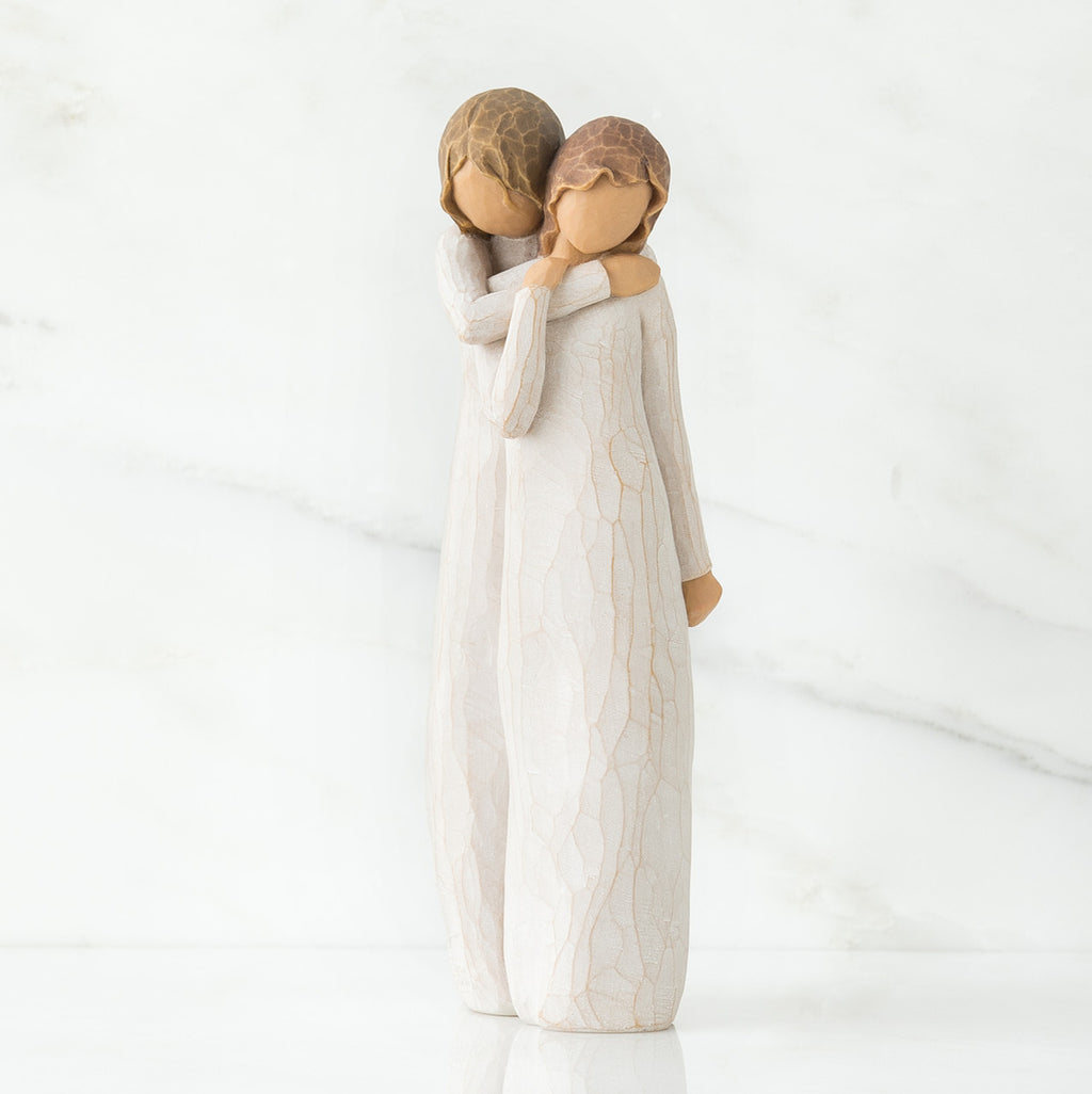 Two faceless woman figurines standing wearing white dresses hugging one another