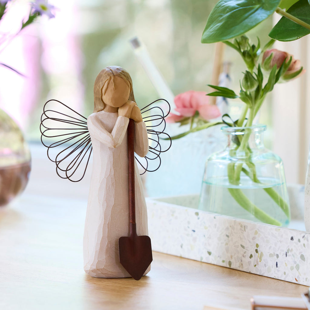 Willow Tree Angel of the Garden, 5.5”h hand-painted standing resin figure in cream dress with wire wings, leaning against brown metal shovel, on table with florals