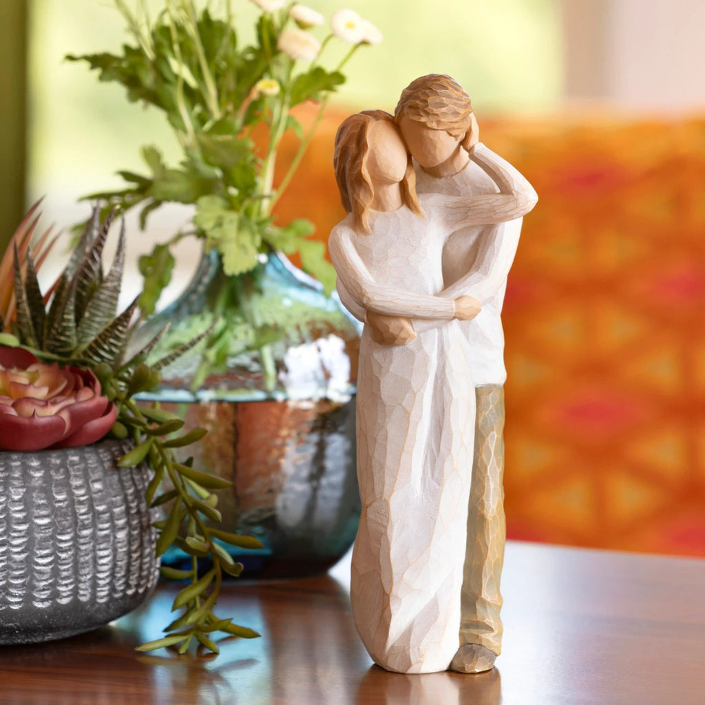 Willow Tree Together, 9”h hand-painted resin figure of couple, standing with arms around each other, dressed in neutral colors, both with lighter skin tones, on table with green florals