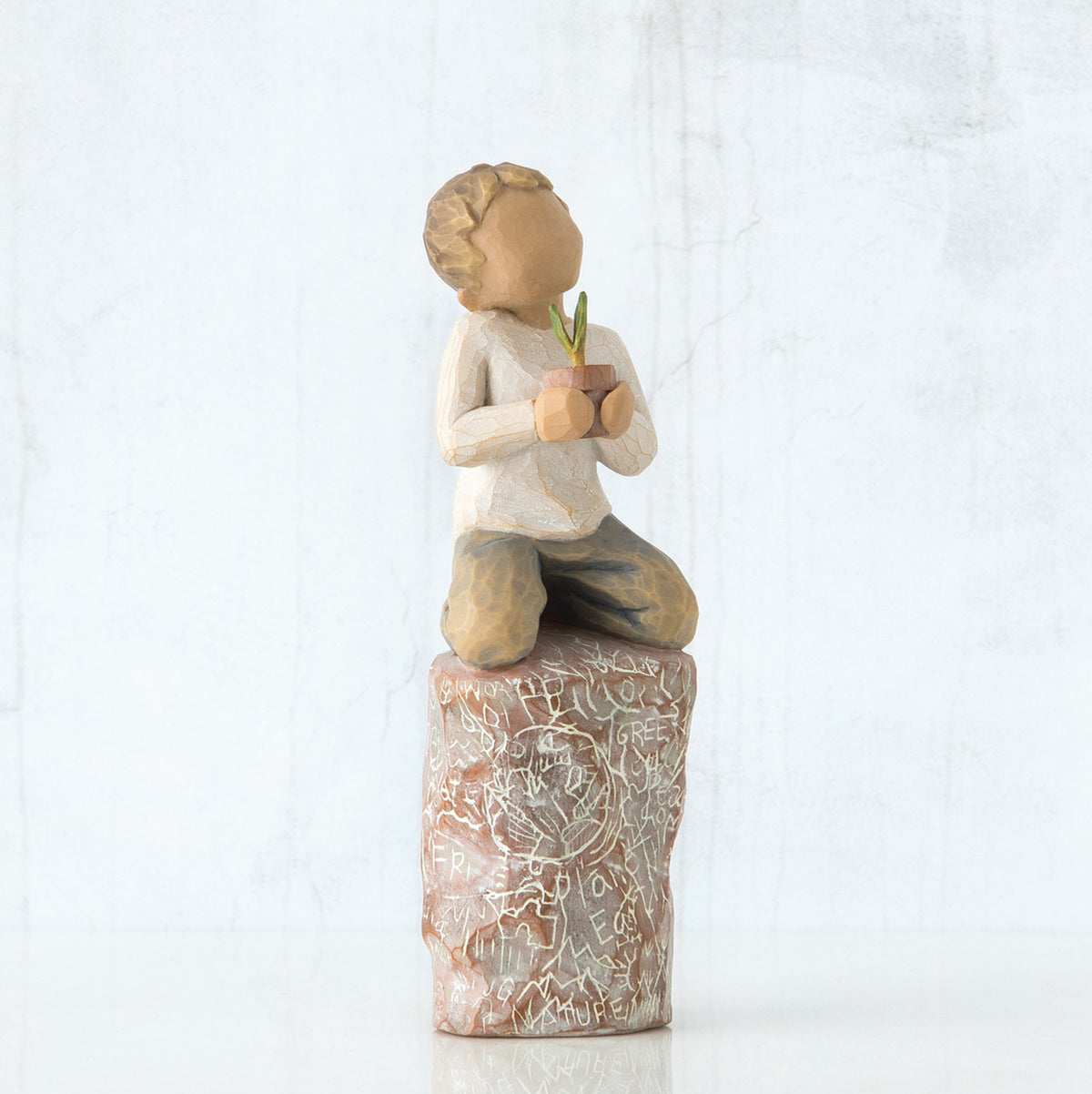 Front view of boy with brown hair sitting on a stump while holding a small potted plant to his chest