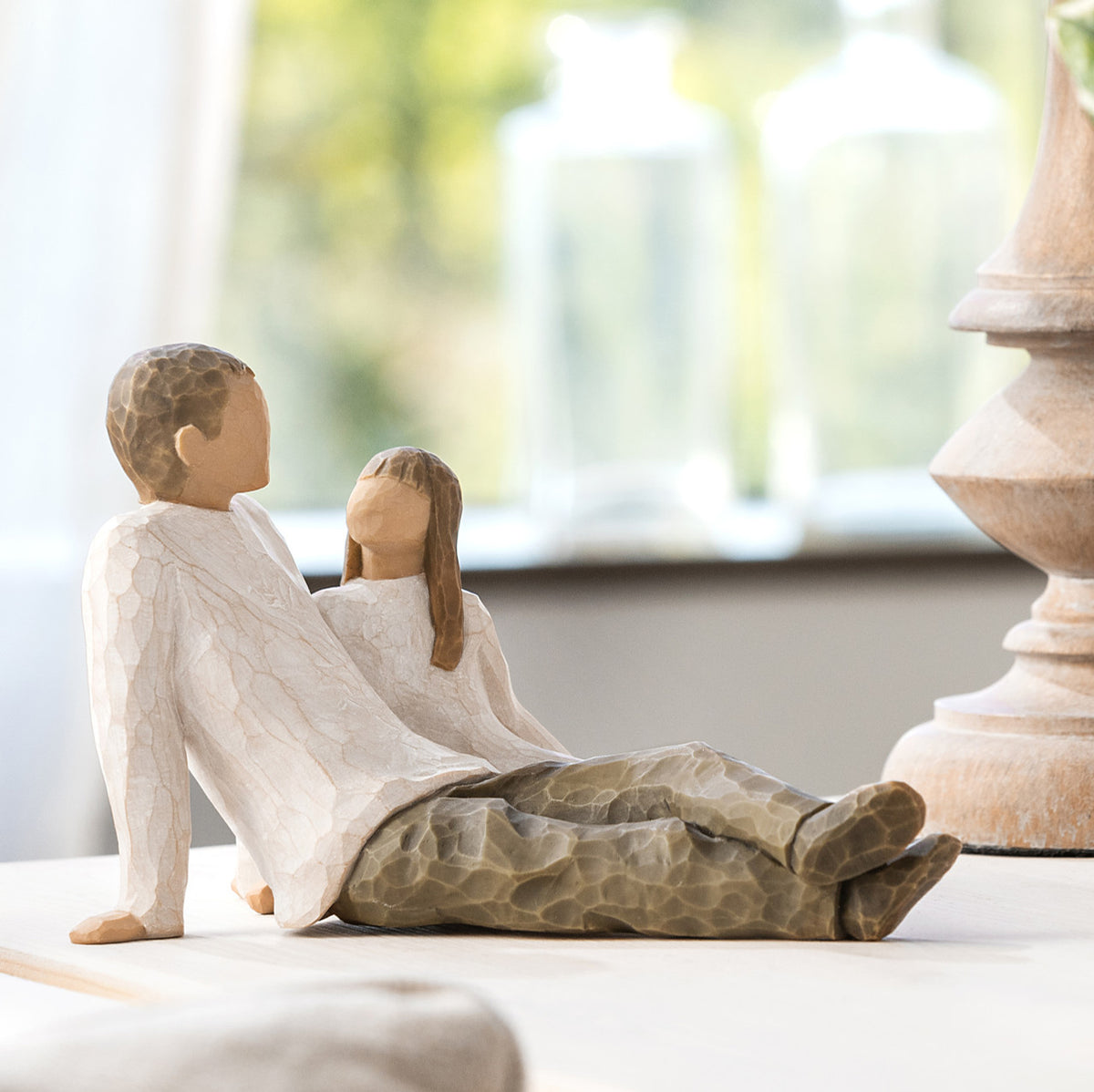 Willow Tree Father and Daughter, 5”h hand-painted resin figure of seated man dressed in neutral colors, legs outstretched, daughter in cream dress by his side, on table by window