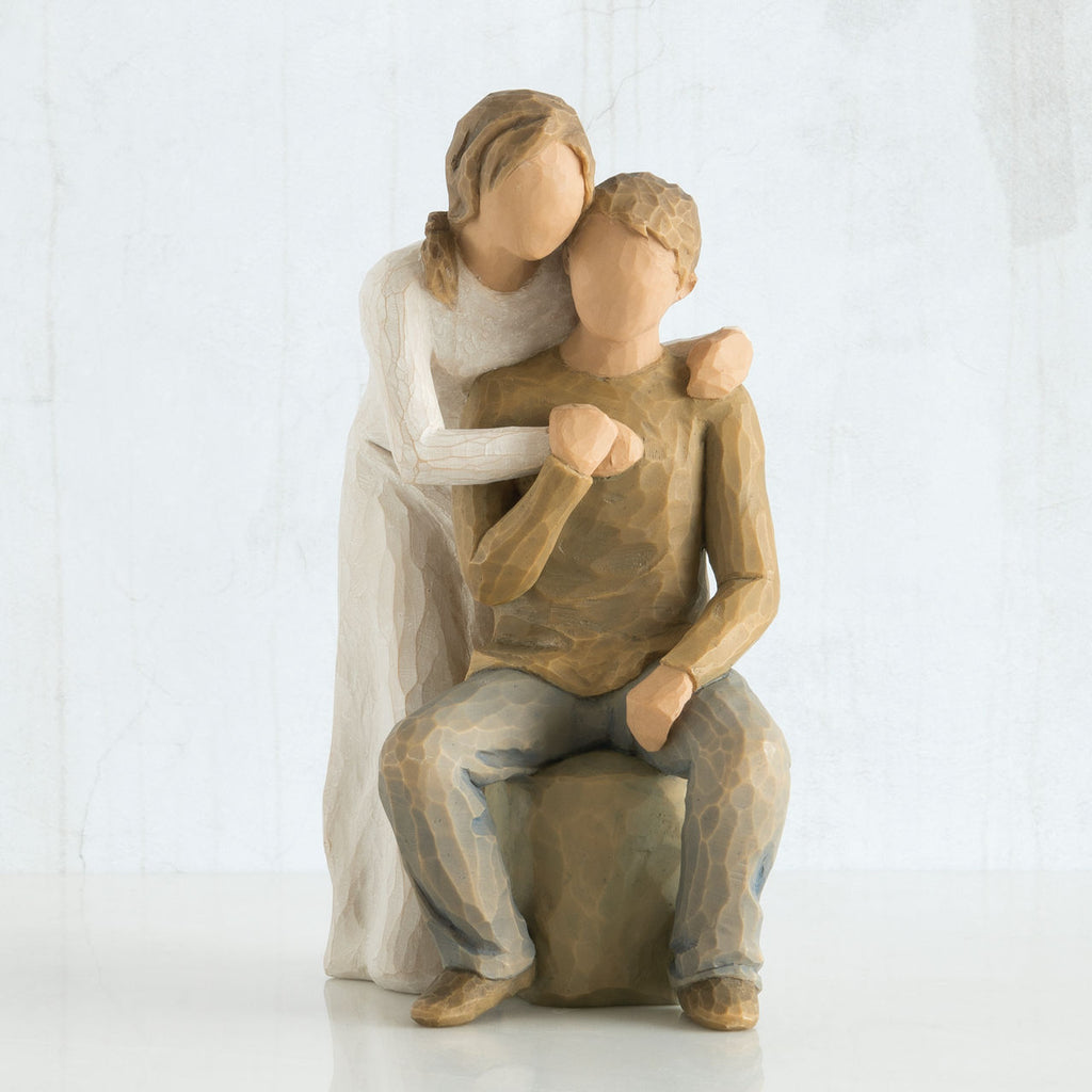Faceless man figurine sitting on rock with faceless woman figurine over his shoulder holding one hand in all white