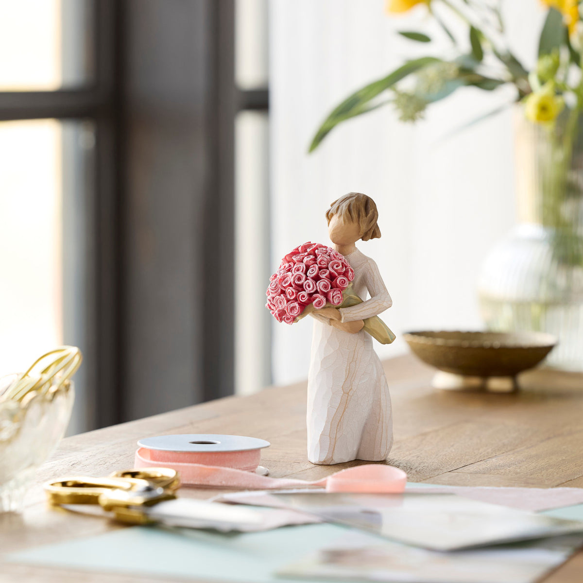 Willow Tree Abundance, 5.5”h hand-painted standing figure in cream dress holding large bouquet of pink roses, on table near window