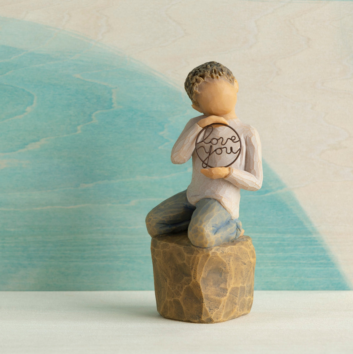 Willow Tree Love you too (lighter skin), 4.5”h hand-painted resin figure of young child in cream shirt and jeans, kneeling on rock, holding wire circle with ‘love you’ inside, in front of blue painting