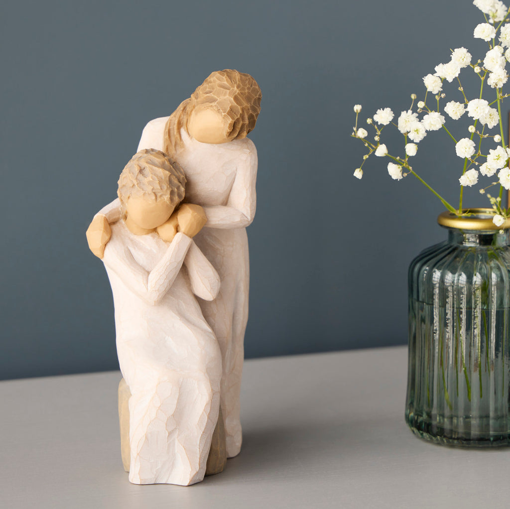 Willow Tree Loving My Mother, 6.5”h hand-painted resin figure of seated older woman with younger woman standing at her shoulder, both in cream dresses, hands clasped, on table with white flowers