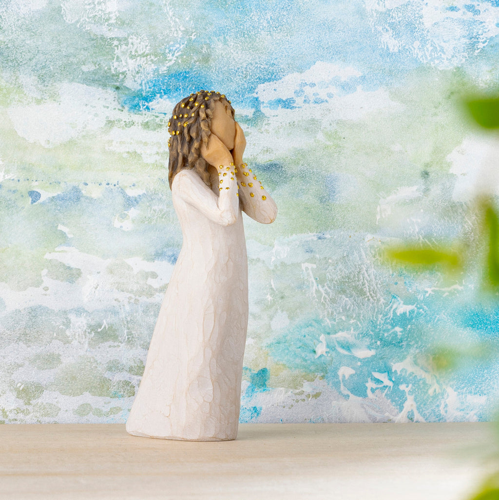 Willow Tree Littlest Christmas Angel, 5.5”h hand-painted standing resin figure in cream dress, both hands up to face in awe, gold leaf dots on sleeves and hair, in front of blue-white painting